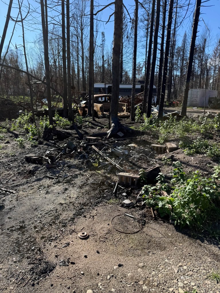 Charred vehicle chassis, scrap metal, and burned timber during wildfire property cleanup
